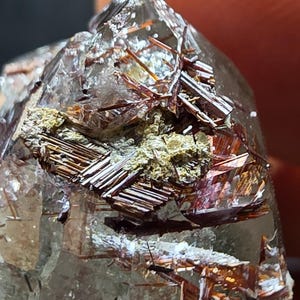 May include: A clear quartz crystal point featuring internal inclusions of reddish-brown rutile needles and a cluster of metallic, bronze-colored hematite plates embedded near the apex of the stone, showcasing natural mineral formations.