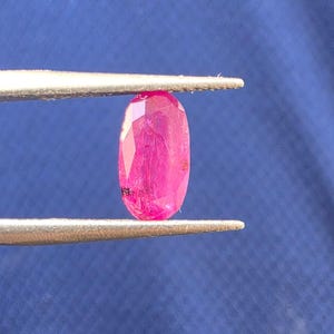 May include: A vibrant, oval-cut pink gemstone held between the metal tips of a pair of tweezers. The stone features a faceted surface that reflects light, displaying internal inclusions and a deep, saturated magenta hue throughout its structure.