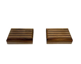 May include: Two wooden coasters with a rectangular shape and a series of horizontal lines. The coasters are made of a light brown wood with darker brown lines, and are set against a white background.