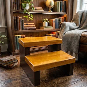 May include: A two-step stool featuring a natural wood finish on the horizontal treads and dark brown vertical supports. The structure is rectangular with a clean design, providing two levels for elevation, constructed from solid wood materials.