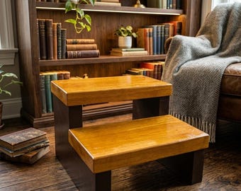 May include: A two-step stool featuring a natural wood finish on the horizontal treads and dark brown vertical supports. The structure is rectangular with a clean design, providing two levels for elevation, constructed from solid wood materials.