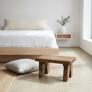 May include: A small, rectangular wooden stool with a dark brown finish and four sturdy, angled legs. The stool features a flat top surface with visible wood grain and is positioned on a smooth, light-colored floor in a room with a bed.