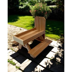 May include: A light-colored wooden picnic table featuring a central rectangular tray area with raised sides, a vertical backrest panel, and two attached benches on opposite sides, constructed from natural wood planks with a visible grain pattern.
