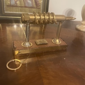 Op de afbeelding: Een goudkleurige metalen sculptuur met een reeks cirkelvormige componenten en een puntig uiteinde, gemonteerd op een rechthoekige houten basis. Een kleine goudkleurige sleutelhanger is bevestigd. De sculptuur wordt tentoongesteld op een gepolijst houten oppervlak.