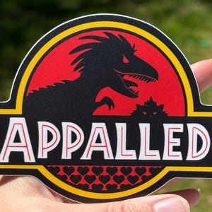May include: A die-cut sticker featuring a circular red background with a black silhouette of a dinosaur and a small cat. The word APPALLED is printed in white capital letters with red outlines across a black horizontal bar with yellow borders.