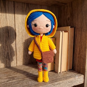 May include: A crocheted doll with blue hair wearing a yellow hooded raincoat, a red shirt, and a brown crossbody bag. The doll has multicoloured patterned leggings and yellow boots. The doll features large black eyes and a light skin tone.