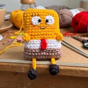 Può includere: Una bambola rettangolare all'uncinetto di colore giallo con grandi occhi bianchi, pupille nere e un piccolo sorriso ricamato. Indossa una sezione di camicia bianca con una cravatta rossa e pantaloni marroni. Gambe gialle e piedi neri.