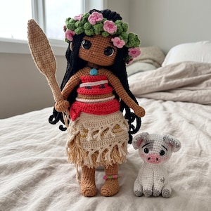 May include: A handcrafted crochet doll with long black hair, a floral crown, and a red and beige outfit, holding a tan crochet spear. Beside the doll is a small white and gray crochet piglet with a pink snout and large black eyes.