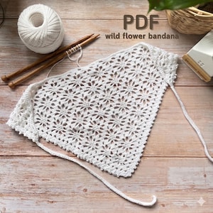 May include: A white, crocheted bandana with a floral pattern, alongside knitting needles and a ball of yarn. The text "PDF wild flower bandana" is visible. The bandana is on a wooden surface.
