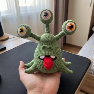 Pode incluir: Um brinquedo de monstro em crochê verde com três hastes, cada uma com um olho redondo no topo. O corpo central apresenta uma boca larga com dentes brancos e uma língua vermelha saliente. A criatura tem várias pequenas pernas pontiagudas.