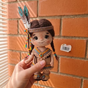 May include: A crocheted doll with brown hair in two braids, wearing a headband with two upright feathers, a tan dress with colorful geometric embroidery, and matching fringed footwear. The doll features large black eyes and rosy cheeks.