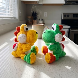 May include: Two handmade crochet dinosaur figures, one yellow and one green, each featuring a white snout, large white eyes with black pupils, and a red spiked ridge along the back. The yellow figure has green feet, while the green figure has orange feet.