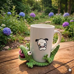 May include: A white ceramic mug with a cartoon dog illustration, held by a green crocheted alligator. The alligator has white eyes and a pink tongue. The mug is on a wooden surface, with blue and purple flowers in the background.