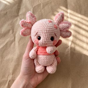Könnte beinhalten: Ein hellrosa gehäkeltes Axolotl-Stofftier mit großem rundem Kopf, markanten äußeren Kiemen, schwarzen Kunststoffaugen und einem korallenfarbenen Shirt. Die ausgestopfte Figur hat eine weiche, texturierte Garnoberfläche mit kleinen rosa Punkten.
