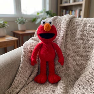 May include: A handmade crochet doll of a red character with a round head, a large orange nose, white eyes with black pupils, and a wide black mouth. The doll has long red arms and legs with textured yarn stitches throughout its entire body.