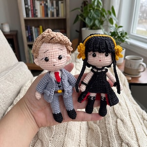 Può includere: Due bambole amigurumi con dettagli all'uncinetto. Una bambola ha capelli castano chiaro, un completo grigio, una cravatta rossa e scarpe nere. L'altra ha capelli neri con accessori floreali gialli, un vestito nero e stivali neri.