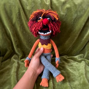 May include: A crocheted doll featuring a wild mane of red and orange yarn, a bright red nose, and a wide open mouth with white teeth. The figure wears a yellow top, a brown collar, and grey trousers, with orange arms and feet made of yarn.