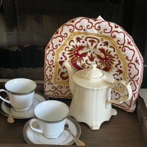 May include: A wooden tray with a white teapot, two white teacups and saucers, and a white tea cozy with a red and gold embroidered design.