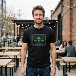 May include: A black short-sleeved t-shirt featuring a graphic of a computer monitor displaying the green text 404 ERROR: BRAIN NOT FOUND. The shirt has a crew neckline and is paired with blue denim jeans, creating a casual look for everyday wear.