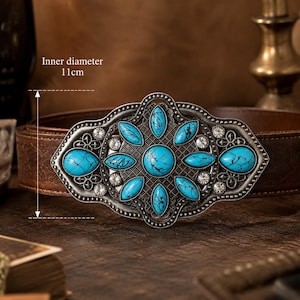 May include: A decorative belt buckle with a silver finish and turquoise-colored stone accents. The buckle has an inner diameter of 4.3 inches and is set on a brown leather belt. The design features a central floral pattern.