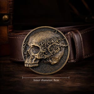 May include: A dark brown leather belt featuring a circular brass-toned buckle with a relief of a human skull adorned with intricate filigree patterns. The buckle has a visible inner diameter of 8 cm, as indicated by the text below the object.