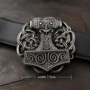 May include: A silver-toned metal belt buckle featuring a detailed Mjolnir hammer design surrounded by intricate knotwork. The buckle is attached to a black leather belt. Text on the image indicates a width of 7.5 cm for the metal component.