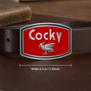 May include: A dark brown leather belt featuring a rectangular metal buckle with a red enamel inlay. The buckle displays the silver-toned text "Cocky" above a detailed silver-toned cockatoo bird graphic. The buckle width is 3.43 inches.