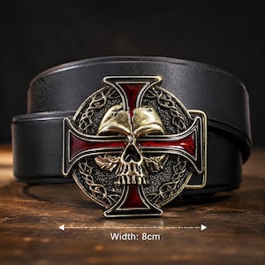 May include: A black leather belt featuring a circular metal buckle with a gold-toned skull centred over a red cross. The buckle design includes intricate filigree detailing around the skull and cross. The buckle has a width of 8 cm.