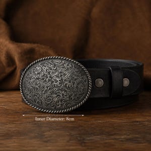 May include: A black leather belt featuring an oval-shaped silver-toned metal buckle with an intricate floral engraving and a twisted rope border. The buckle has a clearly marked inner diameter of 8 cm. The belt strap is secured with two rivets.