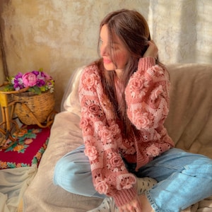 Könnte beinhalten: Ein rosa gehäkelter Cardigan mit 3D-Blumendetails. Der Pullover hat eine Mischung aus hellen und dunklen Rosatönen, wobei die floralen Akzente Textur hinzufügen. Die Ärmel und der Körper sind mit Blumenverzierungen verziert. Der Cardigan wird mit einer blauen Jeans kombiniert.