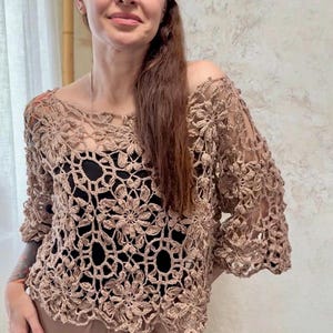 Puede incluir: Una blusa de ganchillo color marrón claro con un intrincado patrón floral y detalles calados. La prenda tiene un escote amplio y mangas tres cuartos, superpuesta sobre una base negra sólida para resaltar el detallado tejido de punto.