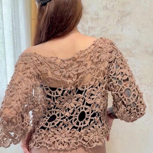 Puede incluir: Una blusa de manga larga tejida a ganchillo en color marrón claro con un intrincado patrón floral y diseño calado. La prenda tiene un escote amplio y se lleva sobre una capa base negra sólida, mostrando una textura detallada tipo encaje.