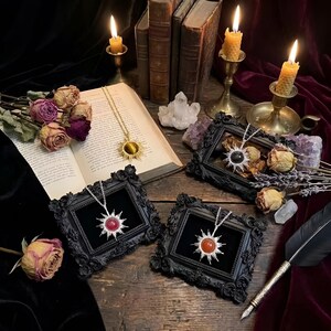 Puede incluir: Una disposición de collares con colgantes en forma de sol, exhibidos en marcos negros ornamentados. Los colgantes presentan varias piedras preciosas, incluyendo ojo de tigre, una piedra negra y piedras rojas y naranjas. La escena está ambientada con velas, libros y flores secas.