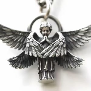 Puede incluir: Un colgante de ángel plateado detallado con alas extendidas y manos juntas. El colgante presenta intrincados detalles de plumas y una pequeña cara de querubín. El ángel está suspendido de una cadena de plata, creando una pieza de joyería religiosa o espiritual.