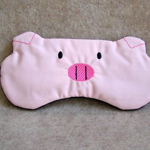 May include: A pink sleep mask shaped like a pig's face, featuring black dot eyes and a pink embroidered snout. The mask has a black border and a tag that reads "An Original MadeByMe".