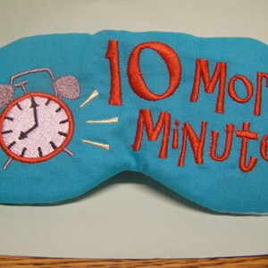May include: Teal sleep mask embroidered with a red alarm clock and the words "10 More Minutes" in red. The alarm clock shows the time as 9:55. The sleep mask is made of a soft fabric.