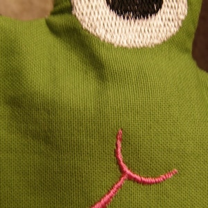 May include: Close-up of a green fabric frog toy with a white and black embroidered eye. The frog has a pink embroidered mouth. The fabric has a textured appearance.