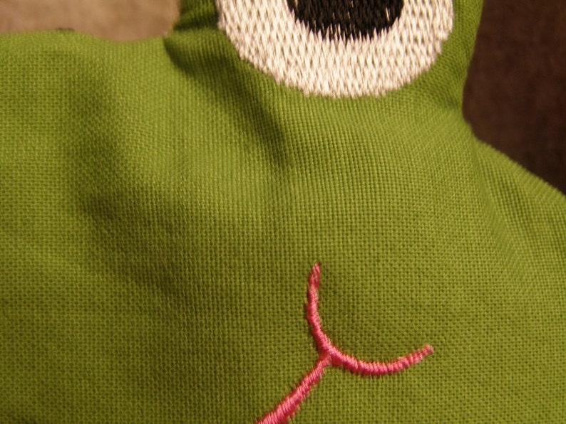 May include: Close-up of a green fabric frog toy with a white and black embroidered eye. The frog has a pink embroidered mouth. The fabric has a textured appearance.