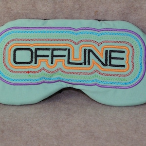 May include: A light blue sleep mask with the word "OFFLINE" embroidered in black. The text is surrounded by colorful stitched lines in orange, purple, green, and red. The mask is designed to block out light.