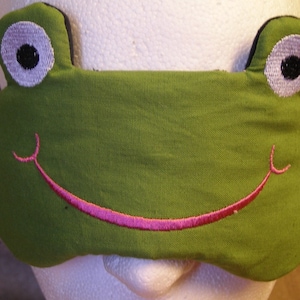May include: A green frog-shaped sleep mask with embroidered details. The mask features large white eyes with black pupils and a pink smiling mouth. The mask is designed to cover the eyes and block out light for sleeping.