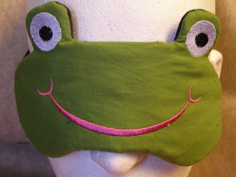 May include: A green frog-shaped sleep mask with embroidered details. The mask features large white eyes with black pupils and a pink smiling mouth. The mask is designed to cover the eyes and block out light for sleeping.