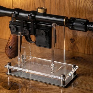 May include: A black blaster prop with a long scope, a wooden grip, and a perforated muzzle, mounted on a clear acrylic display stand. The base of the stand features the engraved text STAR WARS and DL-44 Blaster, with metallic corner accents.
