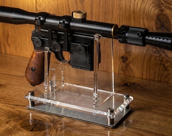 May include: A black blaster prop replica with a wooden grip, a long top-mounted scope, and a muzzle flash suppressor. The blaster is mounted on a clear acrylic display stand featuring the text Star Wars and Blaster DL-44 Heavy Blaster.