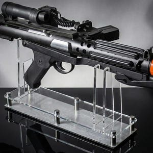 May include: A black sci-fi blaster prop featuring a textured grip, a top-mounted scope, and a coiled black cable. The barrel has a bright orange tip. The blaster is mounted on a clear acrylic display stand with a brushed metal base plate.