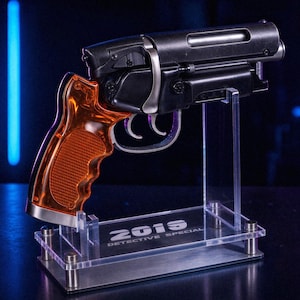 Peut inclure: Un pistolet factice noir et orange avec une poignée orange translucide et une détente argentée, monté sur un support d'exposition en acrylique transparent. La base porte l'inscription blanche 2019 DETECTIVE SPECIAL.