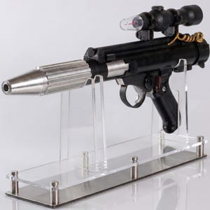 以下が含まれることがあります： 黒と銀色のブラスターピストルの小道具。金属製の円錐形の銃口、黒いハンドル、透明なプラスチック製の照準器を備えた黒いスコープが上部に付いています。アクリル製のディスプレイスタンドと金属製のベースの上に設置されています。