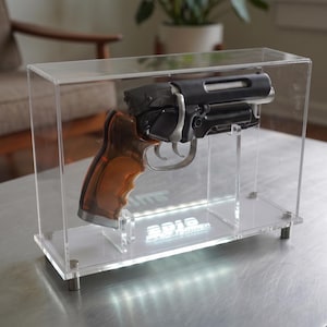 Könnte beinhalten: Eine schwarze und bernsteinfarbene Requisitenpistole in einer rechteckigen Vitrine aus klarem Acryl. Der Sockel der Vitrine zeigt ein weiß beleuchtetes Logo mit der Aufschrift 2019 TARMEN. Die Pistole hat einen silbernen Abzug und einen Griff.
