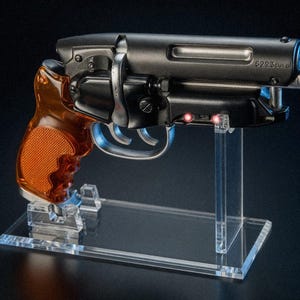 Peut inclure: Un pistolet factice noir mat avec une poignée texturée en ambre translucide et une détente argentée. Le canon porte l'inscription gravée 5223cul.al et deux petites lumières LED rouges sont allumées sur le côté du cadre près du pontet.