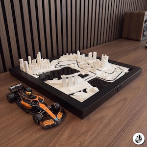 Pode incluir: Um mapa 3D emoldurado em preto do circuito de rua de Fórmula 1 de Singapura com edifícios e marcos em miniatura brancos, acompanhado por um modelo detalhado de carro de corrida laranja e preto com marcas OKX e Google Chrome visíveis.