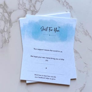 May include: A stack of white cards with the text "Just For You" in elegant script above a blue watercolor wash. The cards include messages of appreciation and encouragement, with a simple design.